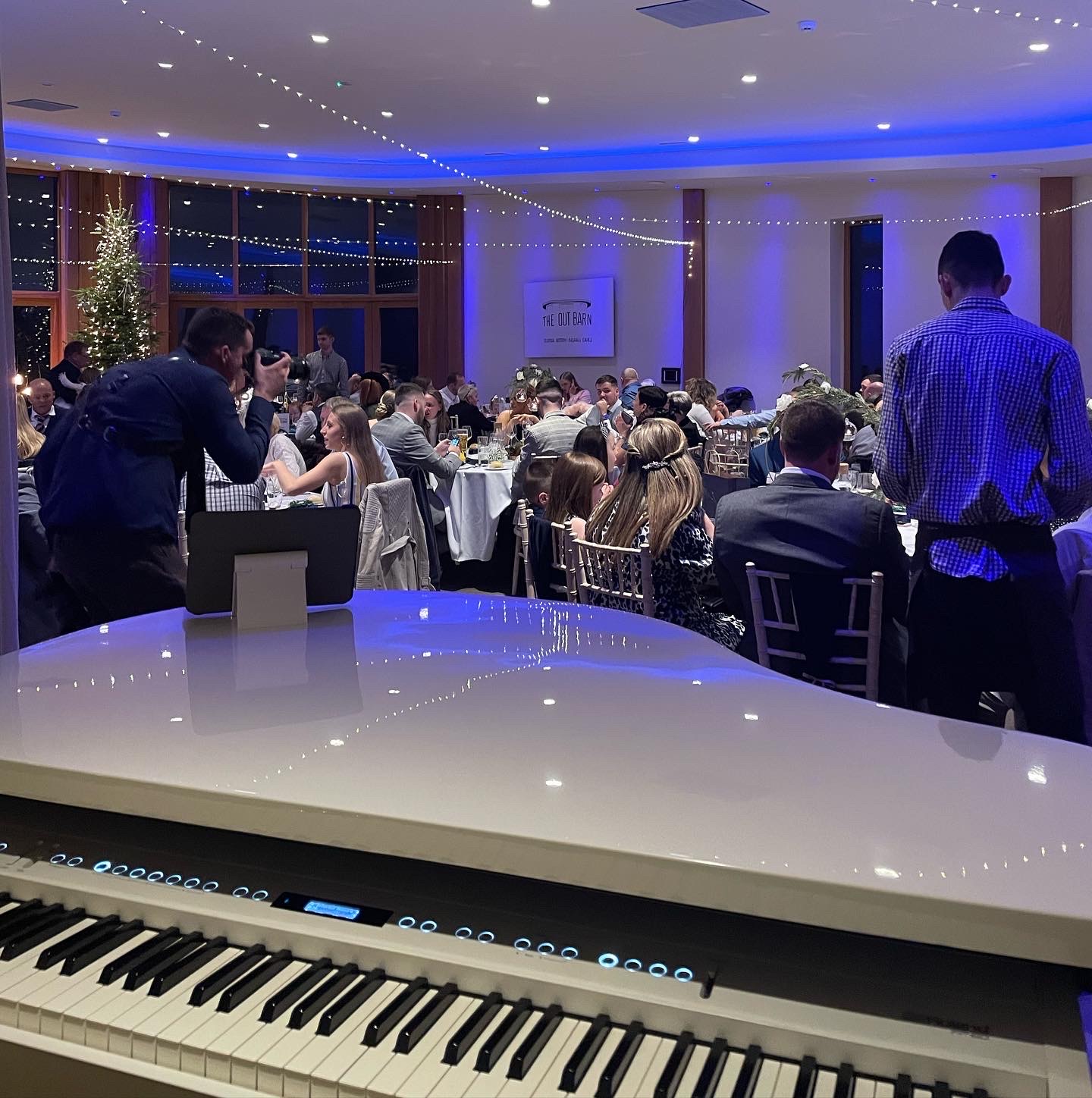 Wedding pianist Craig Smith's white piano reflects stylish purple light during a winter wedding breakfast. The guests are in the background behind the piano and outside everything is dark.